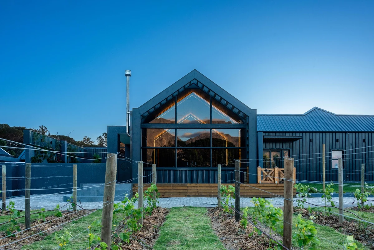 The Glass Castle at dusk: dark charcoal corrugated cladding, a tall steel-framed gable-end wall of glass reflecting the mountains, timber deck and vineyard rows in the foreground at High Riding Country Estate, Sir Lowry's Pass.