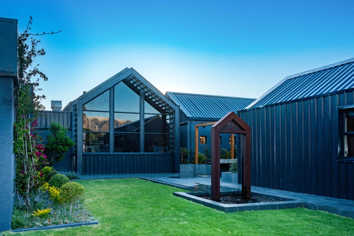The Glass Castle courtyard at dusk: dark charcoal corrugated cladding, tall steel-framed gable-end glazing, a corten-steel water feature and manicured lawn at Sir Lowry's Pass.